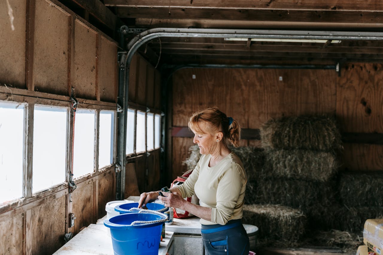 A woman arranging blue buckets in a barn filled with hay bales, standing by a window.